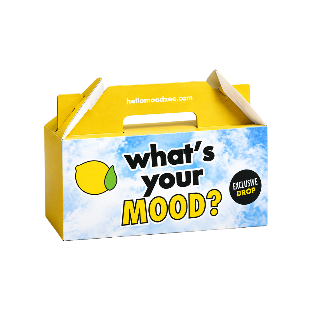 A yellow cardboard product box with a handle, printed with the text 'what's your MOOD?' on a blue sky-and-cloud background, plus a lemon graphic and a black circular 'EXCLUSIVE DROP' badge. The top flap shows the website hellowoodzee.