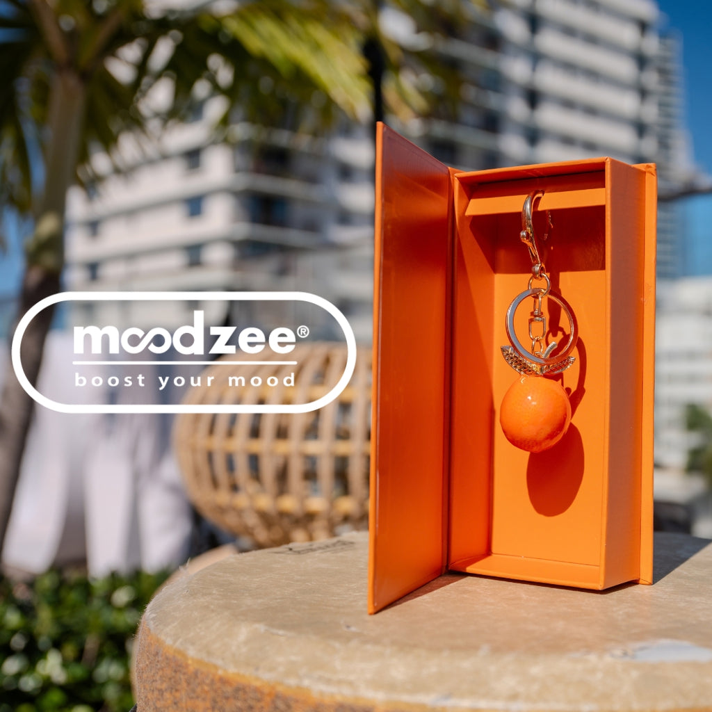 Orange You Glad Moodzee Keychain with an orange fruit charm and gold leaf accent on a gold clasp, displayed in an open orange gift box outdoors in the sun
