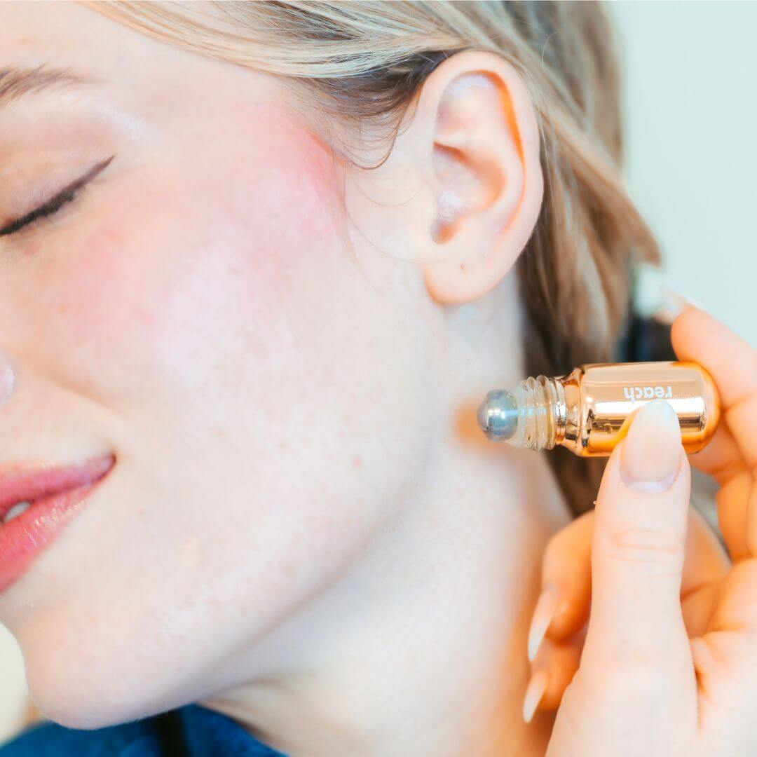 Reach roller ball being applied to pulse point on neck below the ear, gold pill bottle with roller ball tip visible
