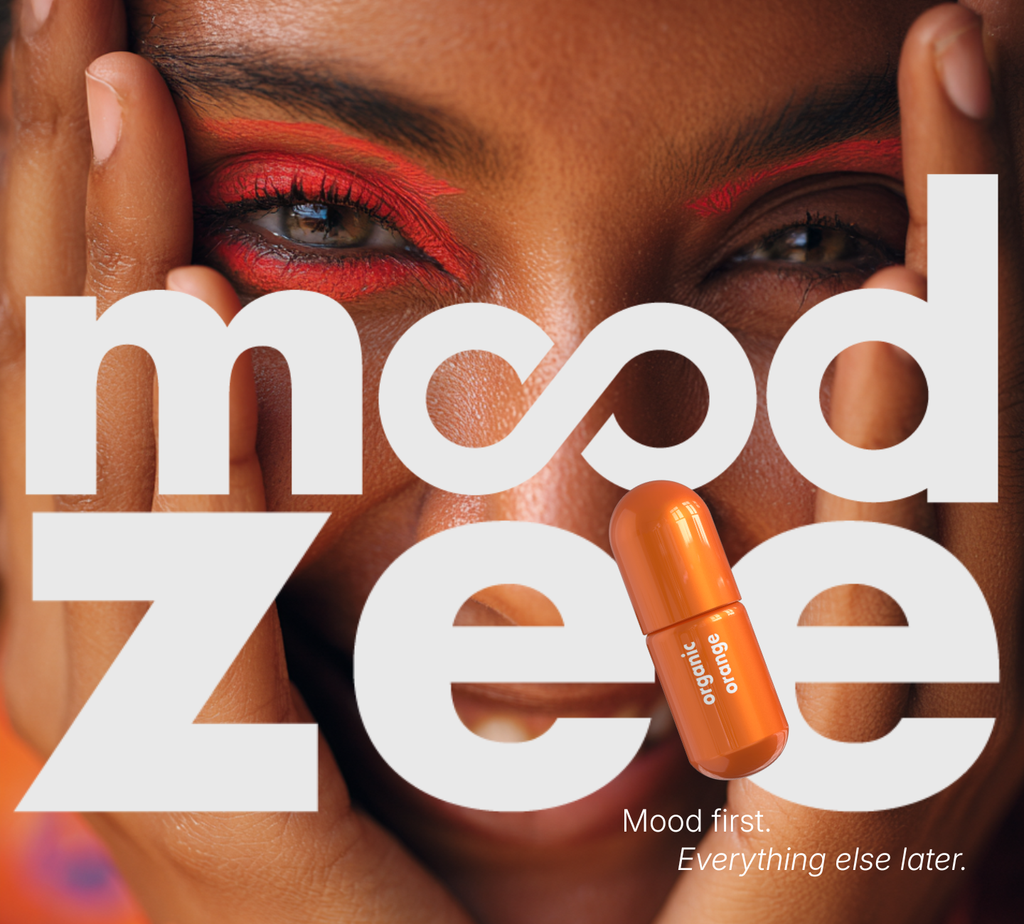 Close-up of a woman with bold red editorial eye makeup holding her face, overlaid with the Moodzee logo and the organic orange essential oil roller ball. Text reads: Mood first. Everything else later.
